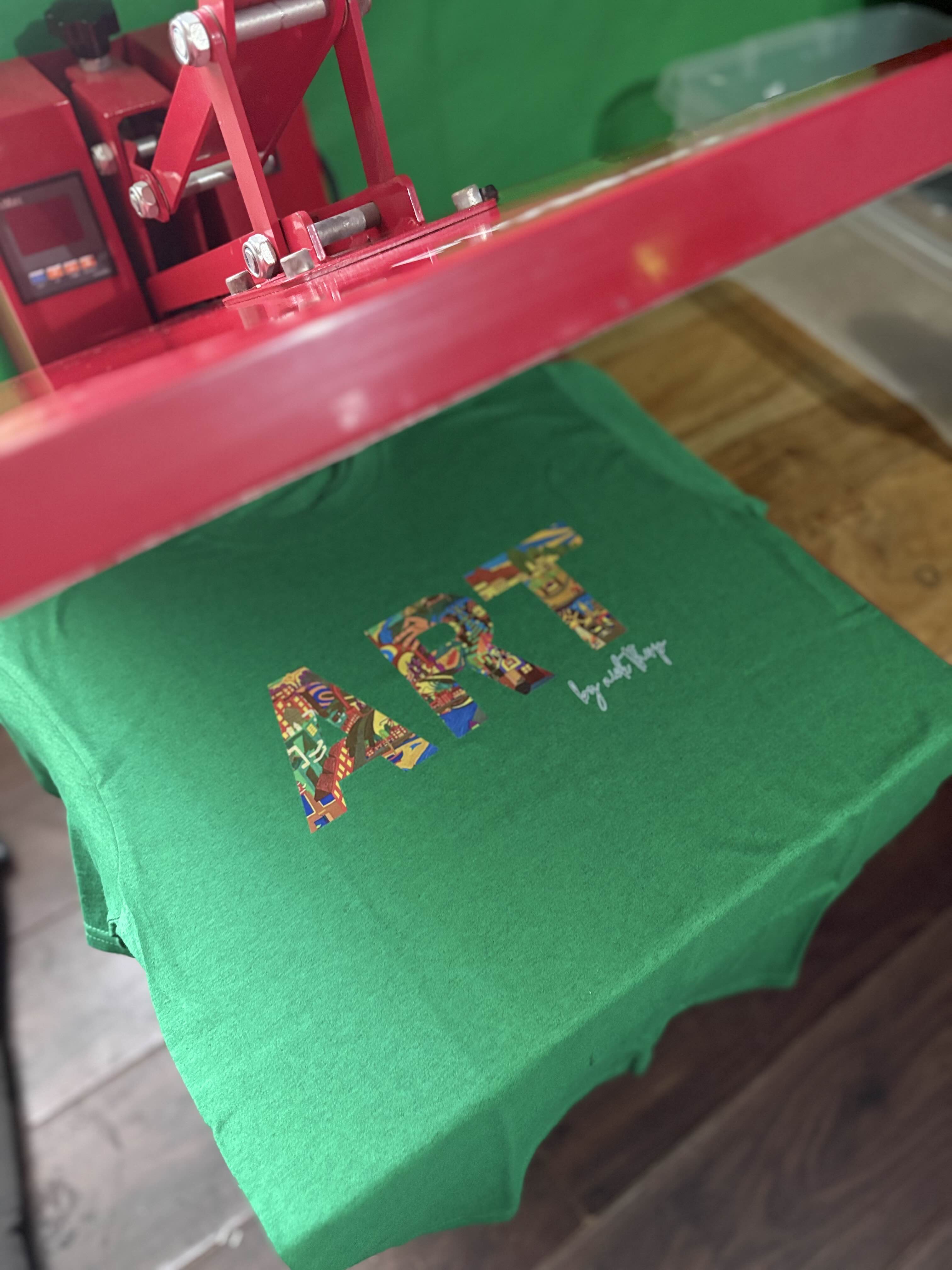 A green t-shirt with a colorful 'ART' graphic and white script text underneath the red press of a heat press machine.