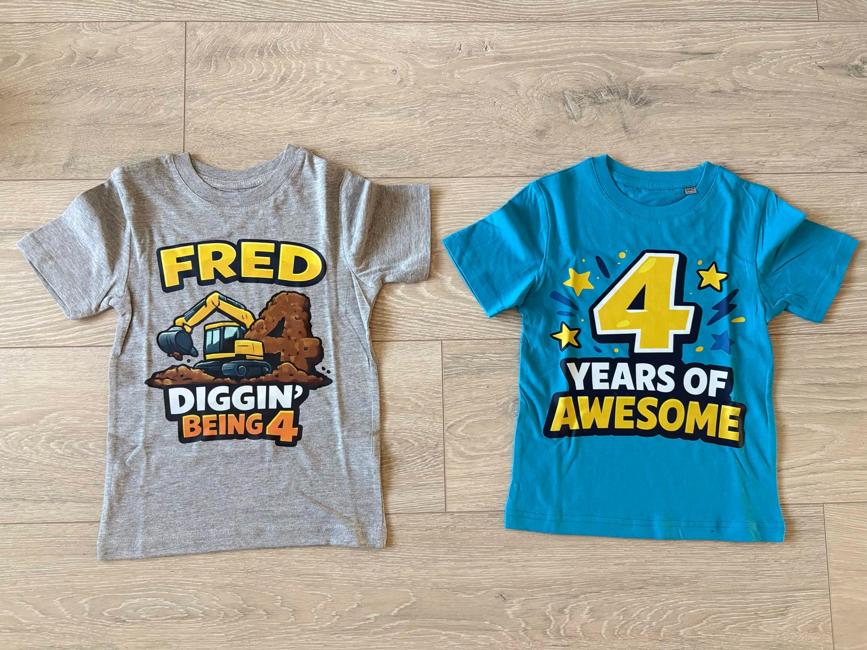 Two children's graphic T-shirts laid flat on a light wood floor. The top shirt is heather gray with a construction graphic reading 'FRED DIGGIN BEING 4' and a yellow excavator.